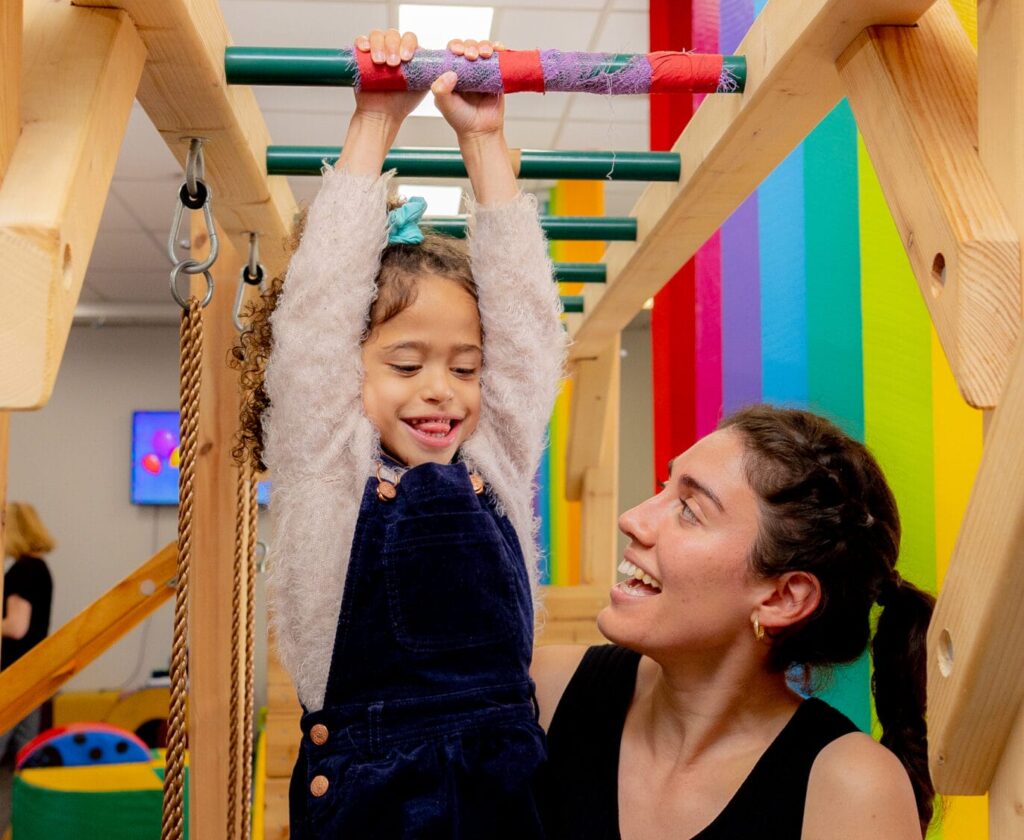 A happy girl hangs from wooden monkey bars, supported by a smiling woman in a colorful playroom.