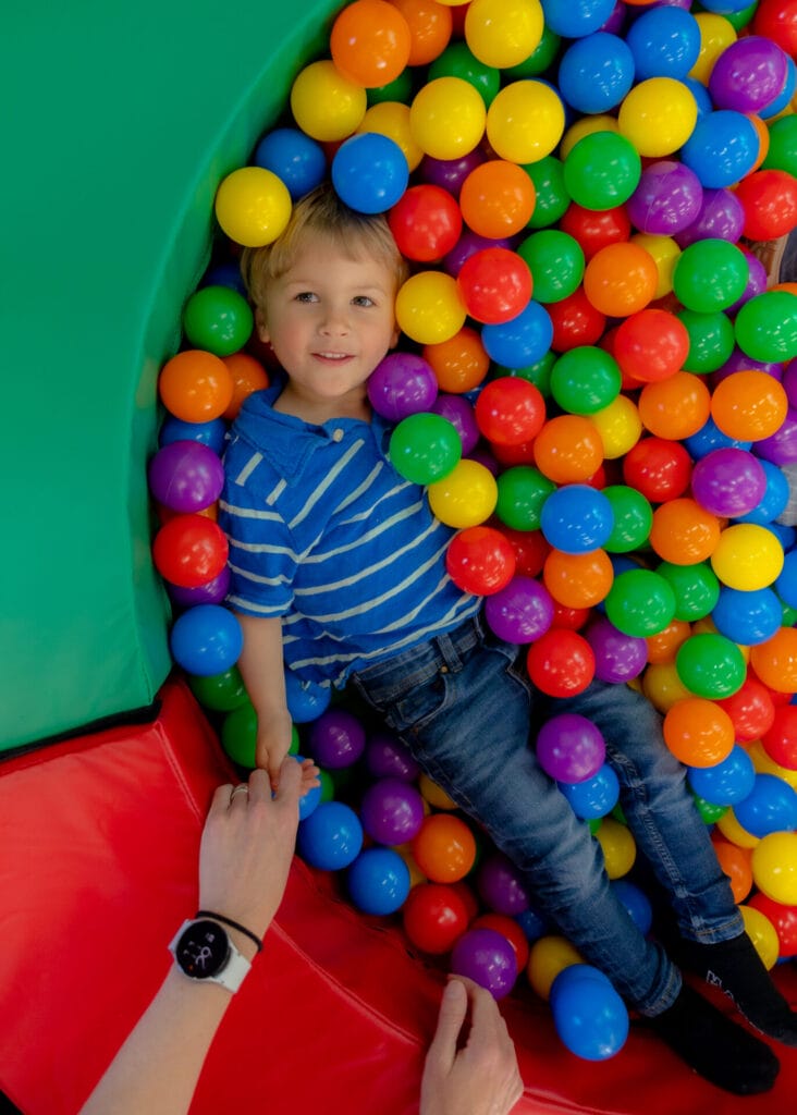 A smiling young boy in a striped shirt lies in a bright, multi-colored ball pit.