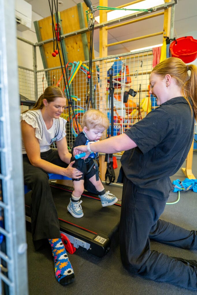 A young child receiving intensive paediatric therapy with two therapists guiding movement on a treadmill.