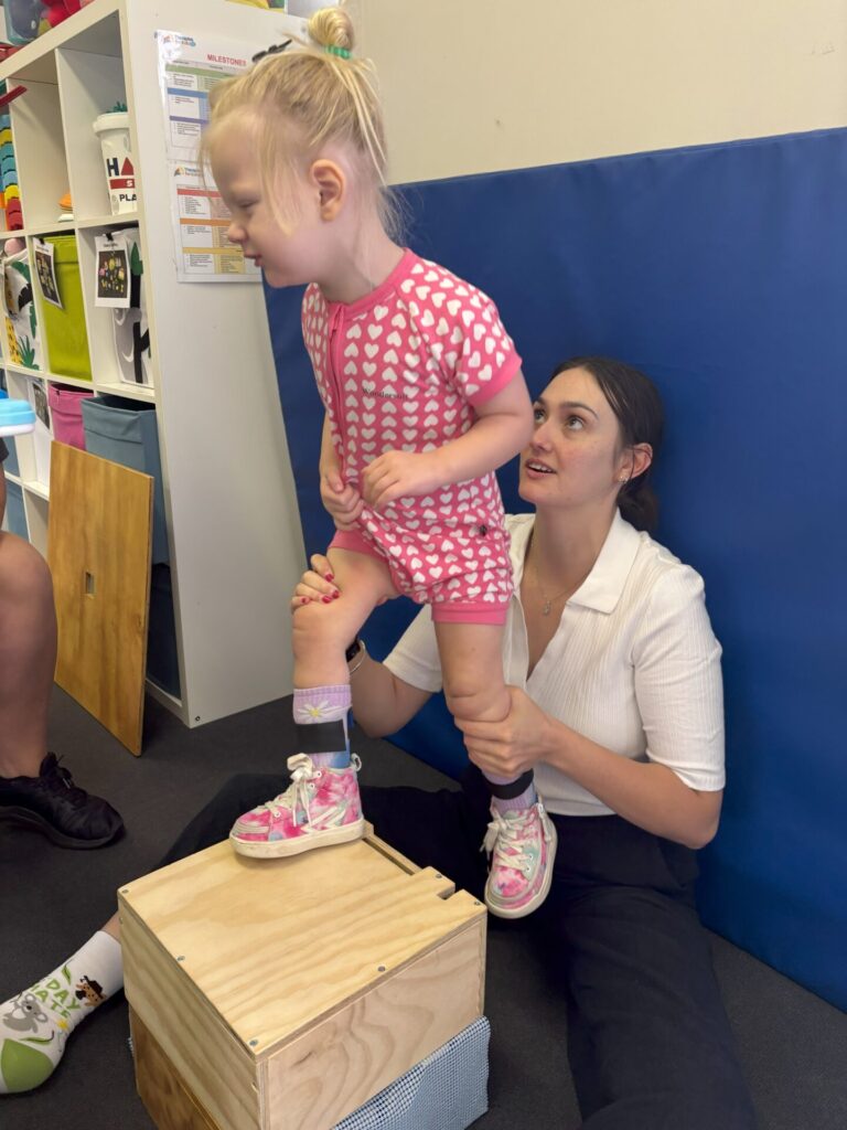 A paediatric therapist supporting a young girl as she practises stepping onto a wooden box during a therapy session at Therapies for Kids.