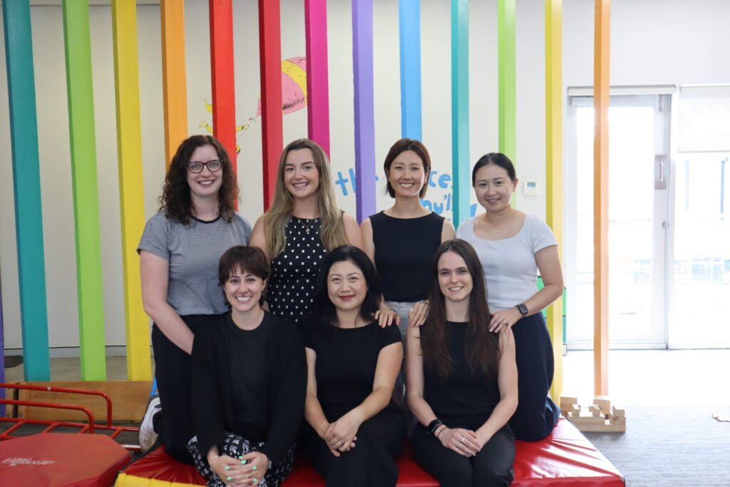 Seven smiling women pose together in front of a bright, rainbow-striped wall.