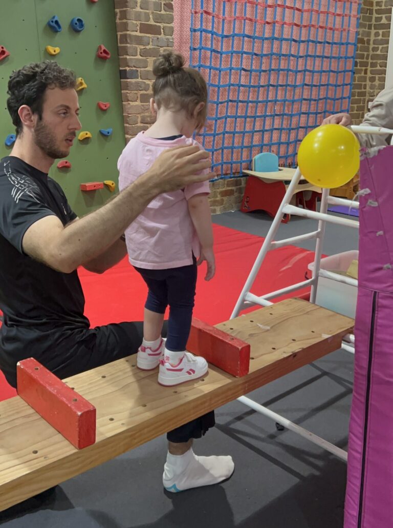 A therapist assists a young girl standing on a wooden balance beam in an indoor sensory gym.