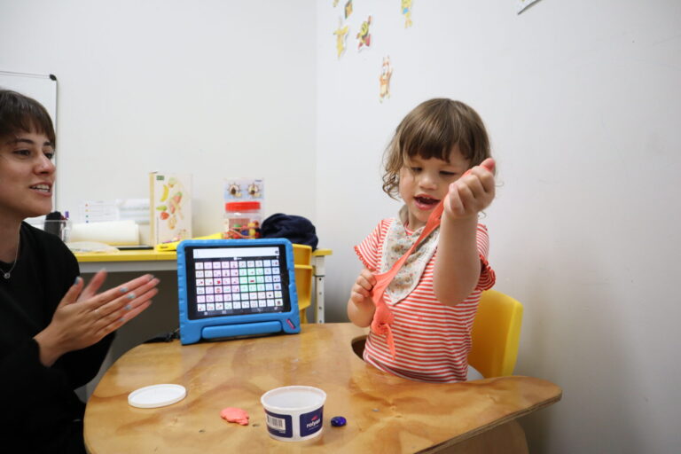 A child stretches pink putty at a table next to a communication tablet while an adult claps in encouragement.