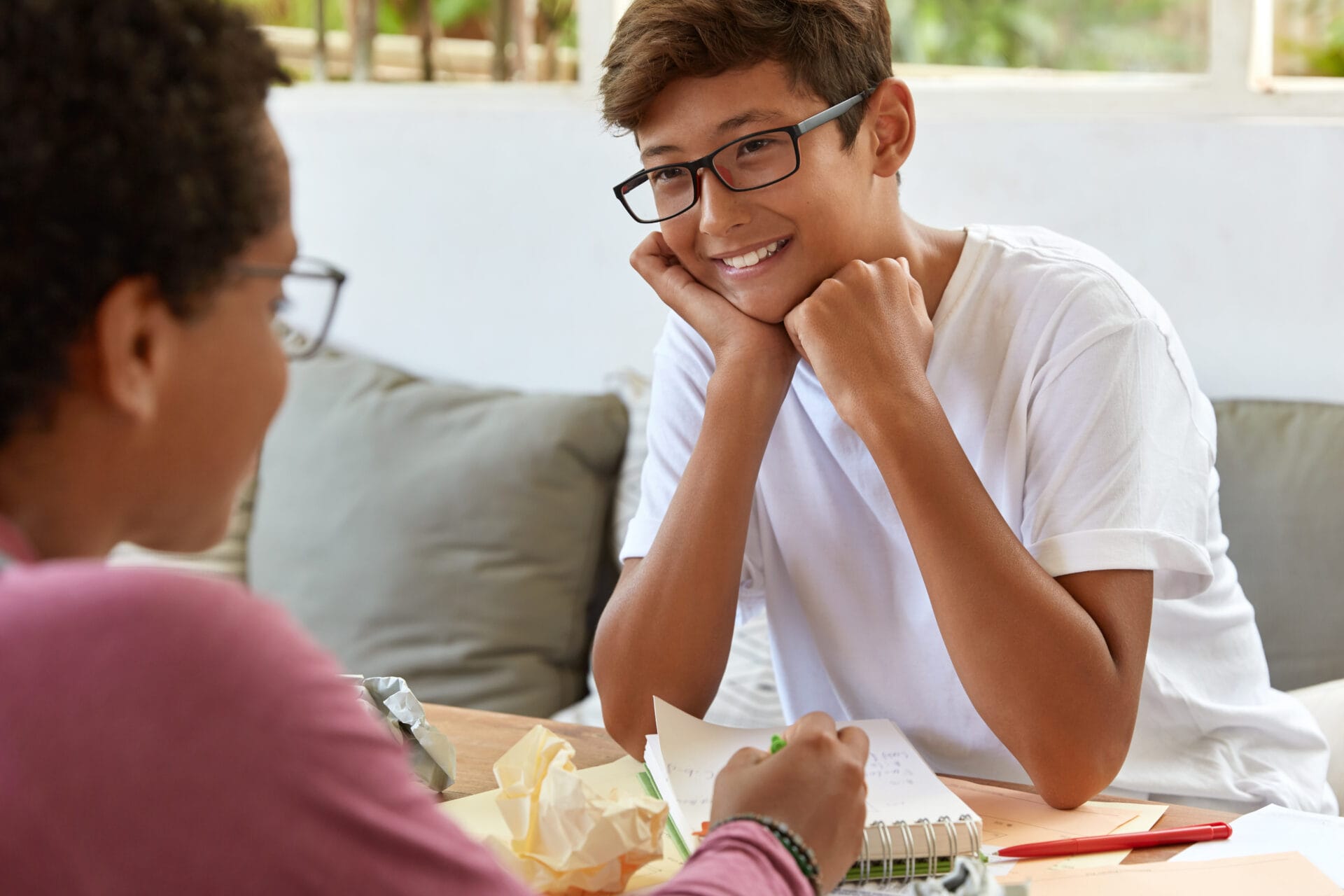 Happy male teenager dressed in white casual t-shit, looking at a tutor who is pointing to a notepad with writing.