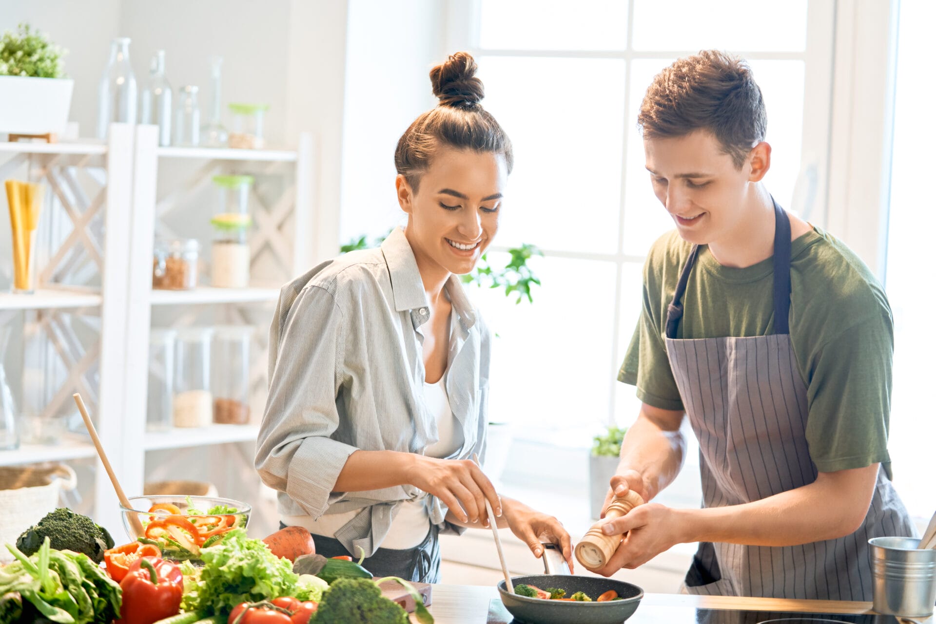 Smiling young man and woman cooking together in a bright kitchen, with the man grinding pepper and woman stirring a pan.