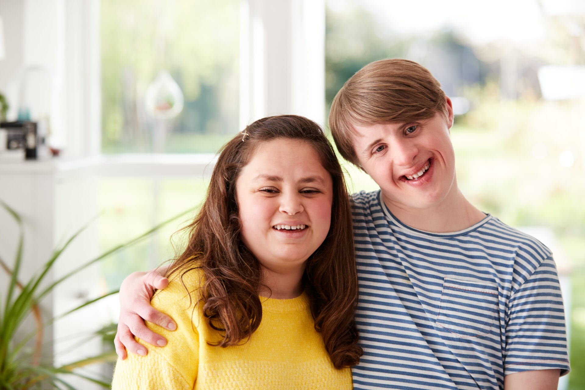 Smiling young woman in a yellow sweater and young man in a striped shirt, both with Down syndrome, embracing.