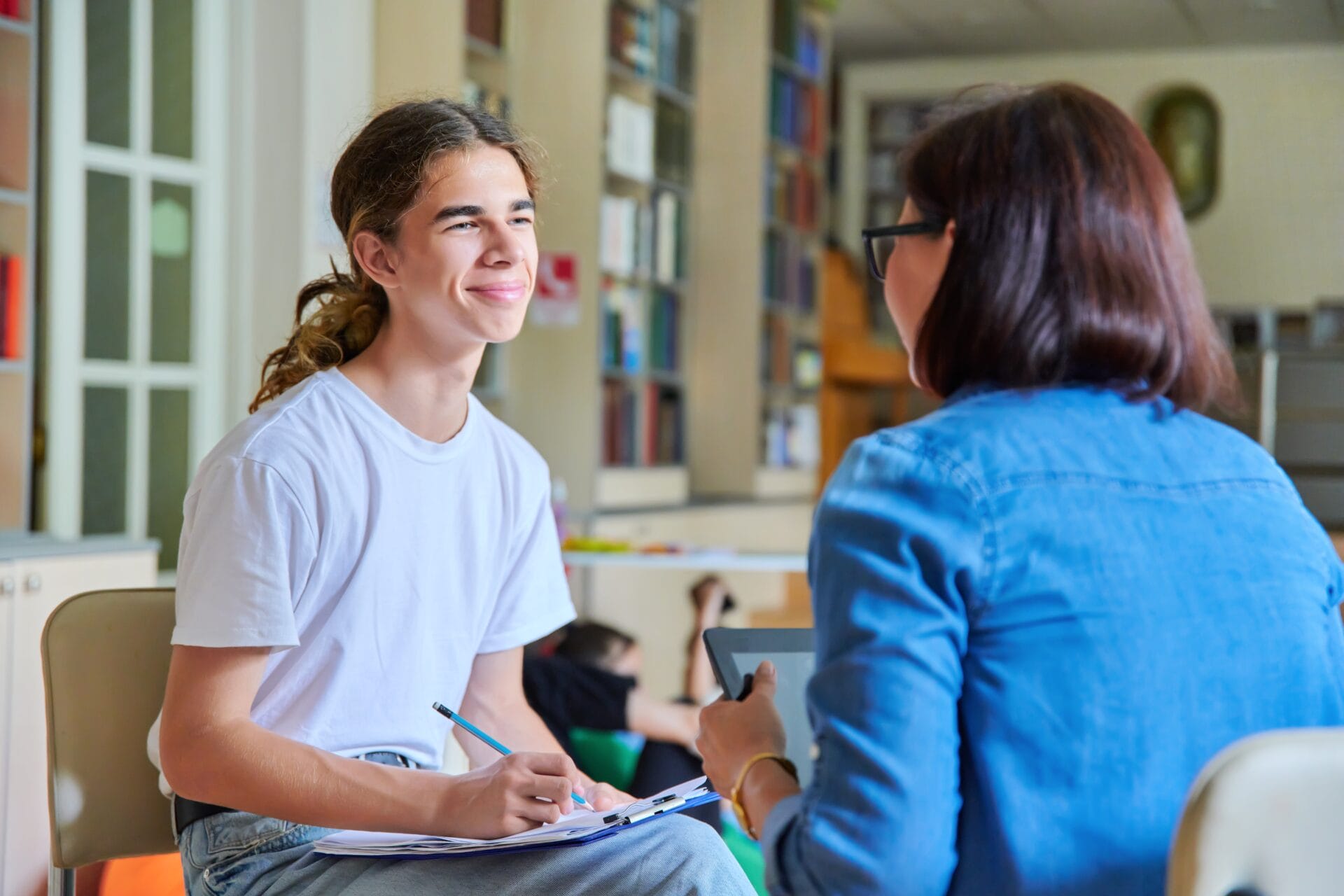 Woman school psychologist teacher talking and helping student, male teenager, inside a library.