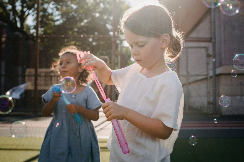Two young children blowing bubbles outdoors in the late afternoon sun, one in the foreground holding a pink bubble wand.