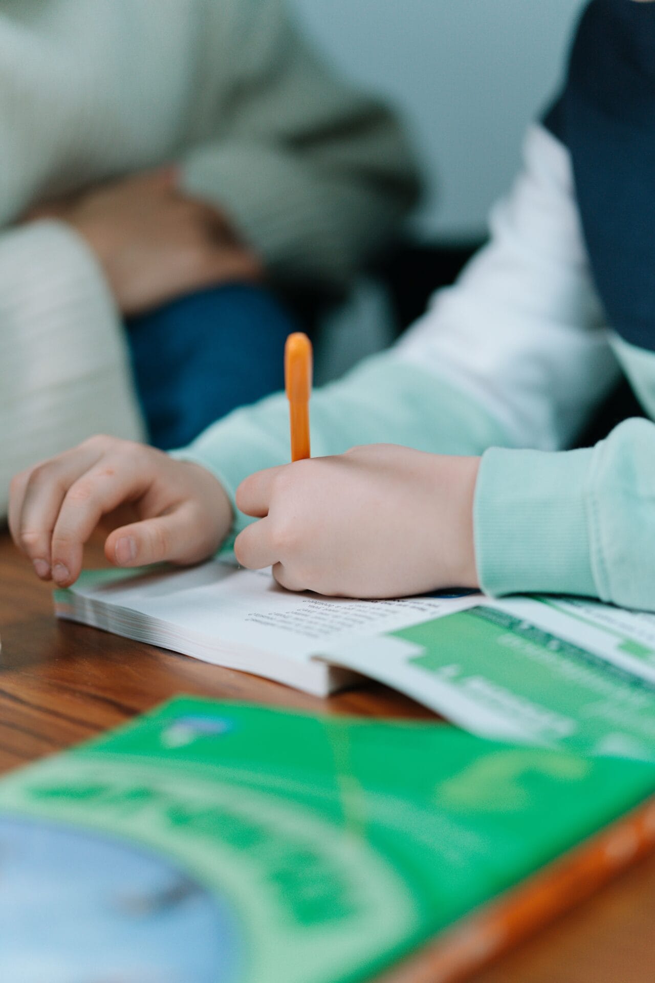 Close-up of a child’s hands holding an orange pencil and writing in a workbook.