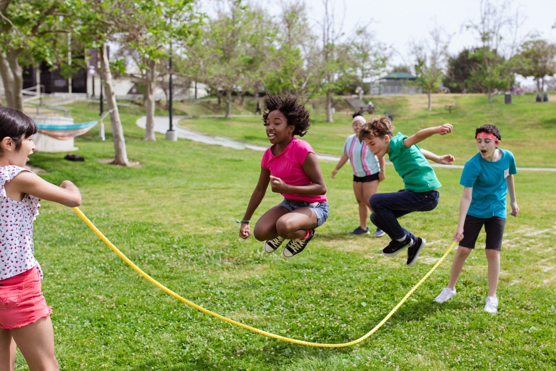 A group of children taking turns jumping over a long skipping rope in a park.